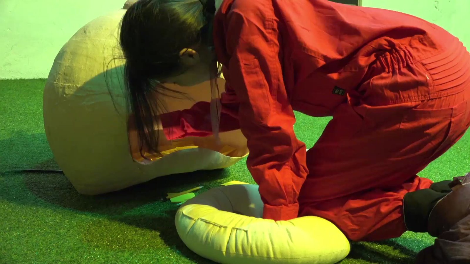 Performance-art photograph: a person in a red jumpsuit kneeling on green turf among large soft white sculptural forms under green light