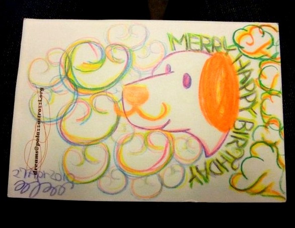 Colorful crayon drawing of a curly, lion-like dog character in orange and pastel swirls, with the handwritten words 'Happy Birthday'